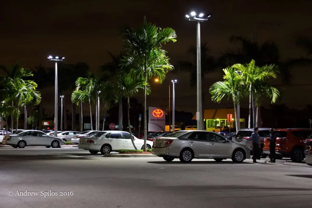 A brightly lit car dealership parking lot at night with palm trees, illustrating factors that impact parking lot light replacement cost.