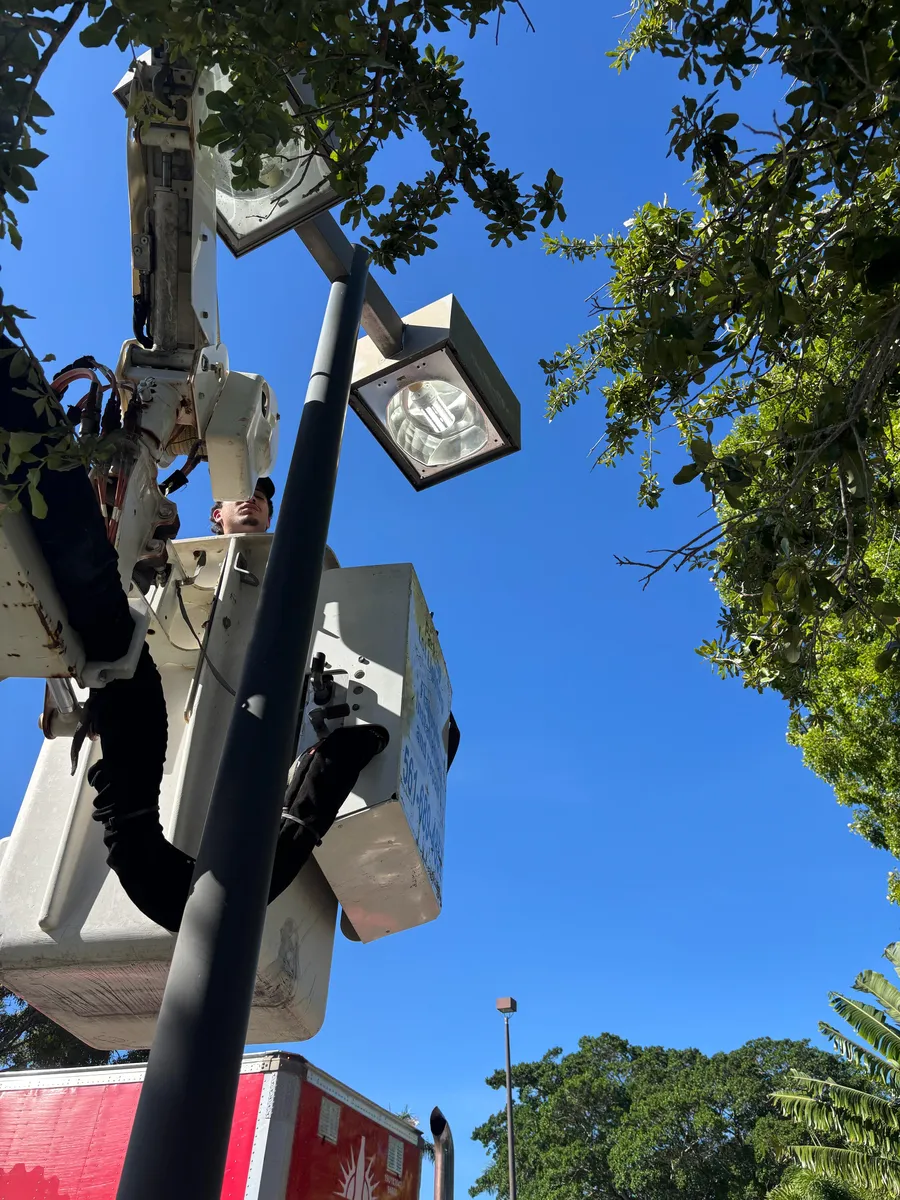 Low Angle View Of A Technician From American Lighting And Electrical Services In A Bucket Truck Working On A Pole-Mounted Light Fixture During The Day.